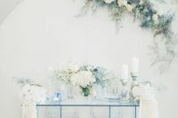 a white backdrop decorated with white and serenity blue blooms and an ice blue sheer glass table and blooms for an ethereal look