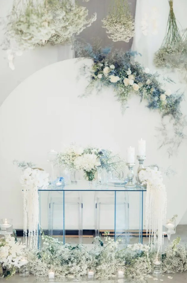 a white backdrop decorated with white and serenity blue blooms and an ice blue sheer glass table and blooms for an ethereal look