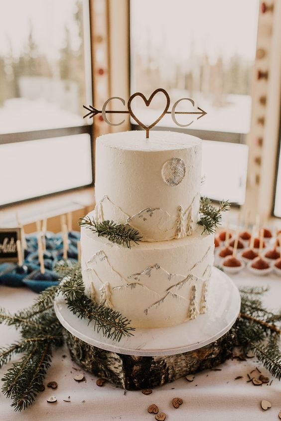 a white buttercream wedding cake with mountains, fir branches and a cute and cool topper with letters and a heart