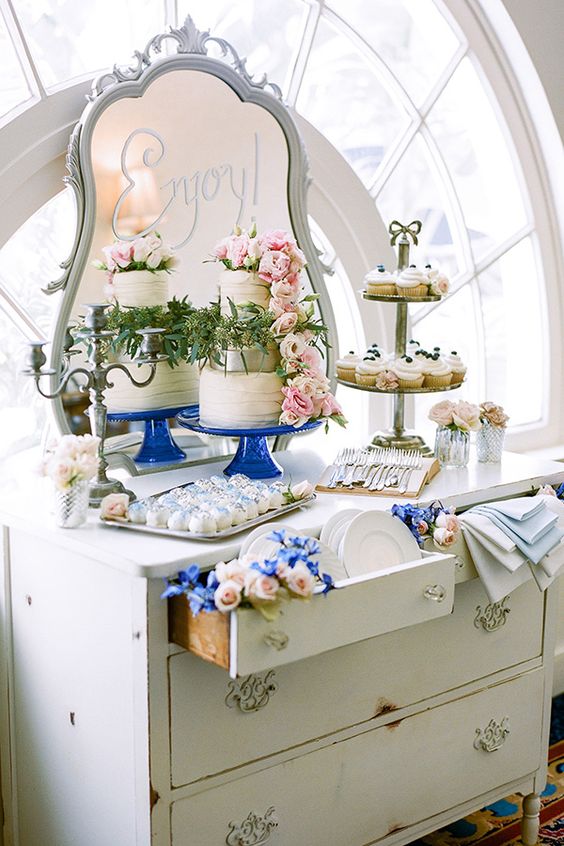 a white vintage dresser as a wedding dessert table, with pink and blue blooms, white and blue napkins, a wedding cake with pink blooms and desserts
