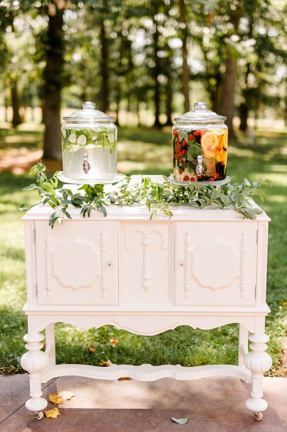 a white vintage dresser as a wedding drink station, with a greenery runner and drinks in tanks is an amazing idea