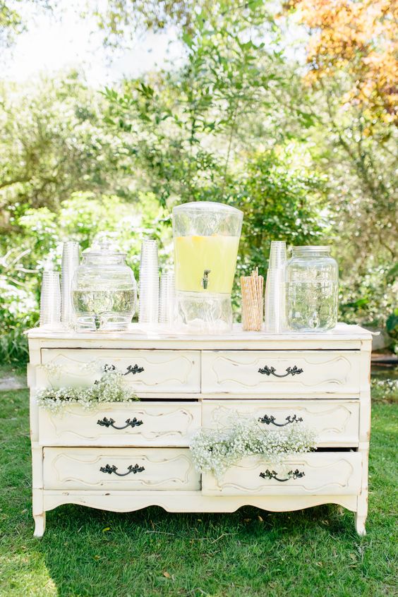 a white vintage dresser decorated with baby's breath and with lemonade and water tanks as a wedding drink station