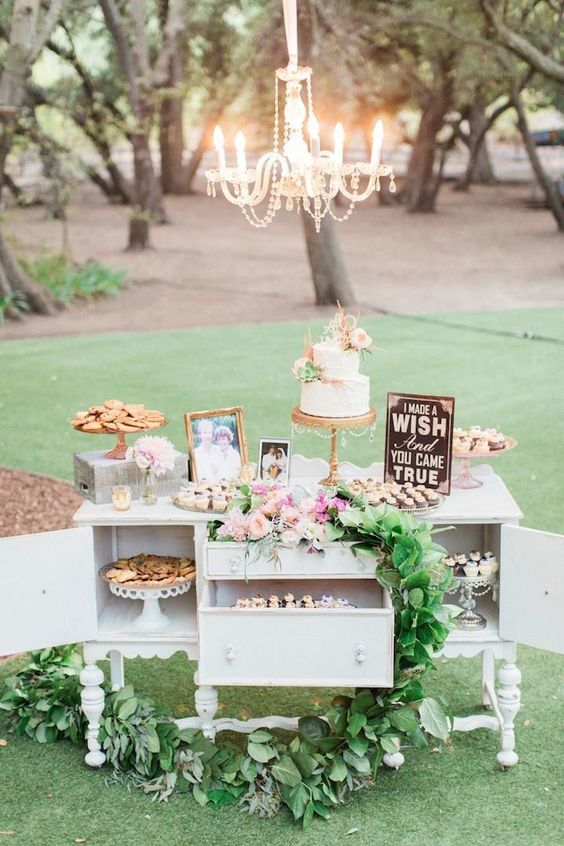 a white vintage dresser decorated with pink blooms and greenery, a chalkboard sign and photos, cookies and wedding desserts of various kinds