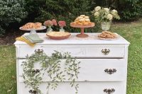 a white vintage dresser with greenery and a white floral arrangement as a wedding dessert table,w ith lots of delicious desserts