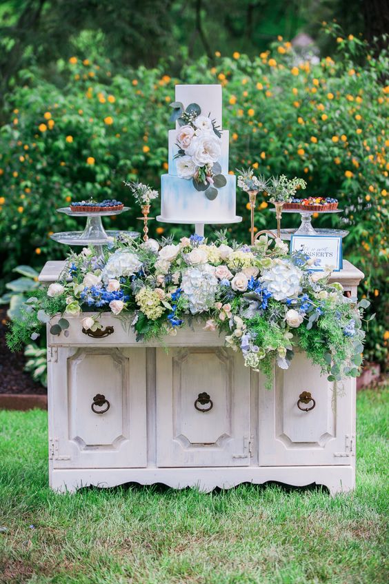 a white vintage dresser with neutral and pastel blooms and greenery various delicious wedding desserts for a vintage wedding