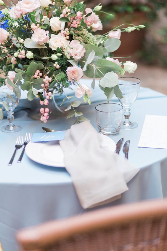 an airy wedding tablescape with a blue tablecloth and neutral napkins, blush blooms and berries plus greenery