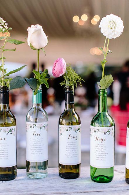 an assortment of wine bottles with blooms and greenery and a seating chart on them is a very creative idea for a vineyard wedding