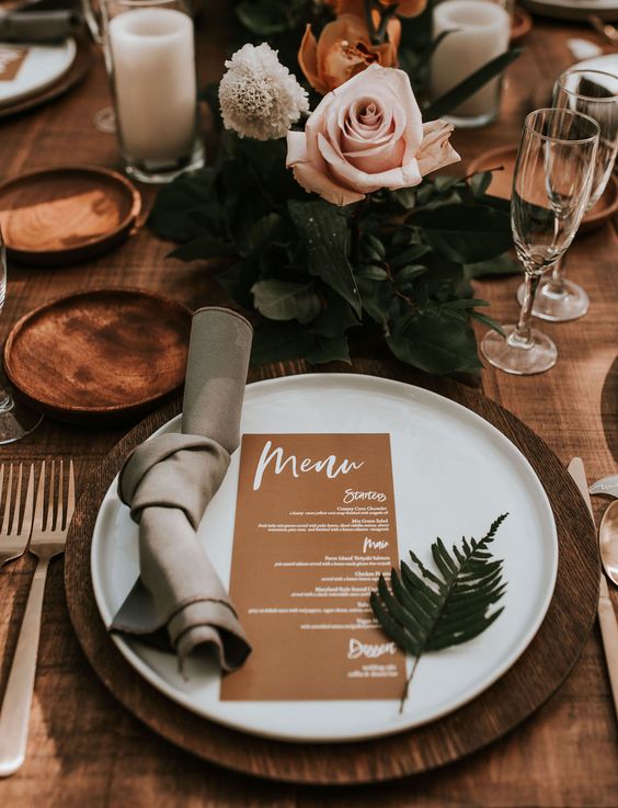 an elegant and chic wedding tablescape with wooden chargers, white plates, delicate blush and rust blooms, a kraft menu and greenery
