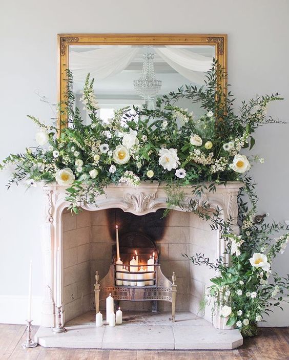an exquisite fireplace with candles inside it, a vintage framed mirror and lush greenery and white blooms