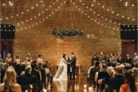 an industrial wedding ceremony space with lights over the space, candles to line up the aisle and a greenery chandelier