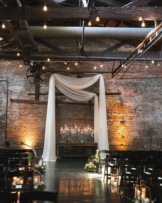 an industrial wedding space decorated with white curtains, candles in glass candleholders, candles to line up the aisle, greenery on the chairs