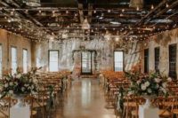 an industrial wedding venue decorated with lights, with lush floral and greenery arrangements and greenery on chairs