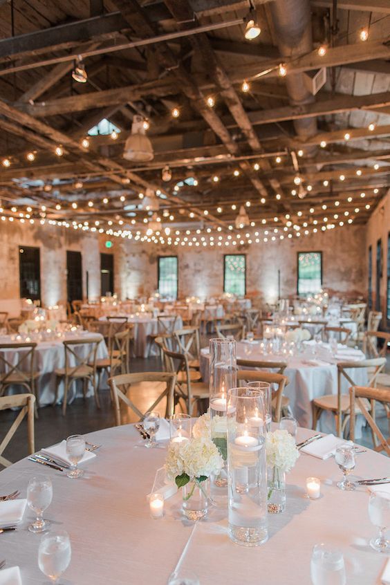 an industrial wedding venue with brick walls, wooden beams, hanging lights, lamps and white linens and blooms