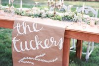 an outdoor wedding reception table with a kraft paper runner with calligraphy, greenery and tall and thin candles