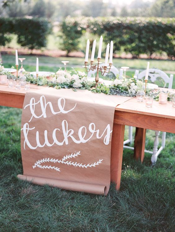 an outdoor wedding reception table with a kraft paper runner with calligraphy, greenery and tall and thin candles