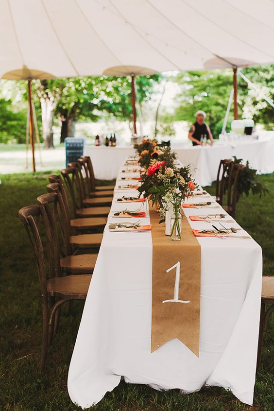 an outdoor wedding reception table with a white tablecloth and a kraft paper runner with a number, bold florals and ombre napkins