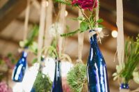 blue wine bottles with greenery and blooms hanging on burlap are lovely decorations for a rustic or woodland wedding