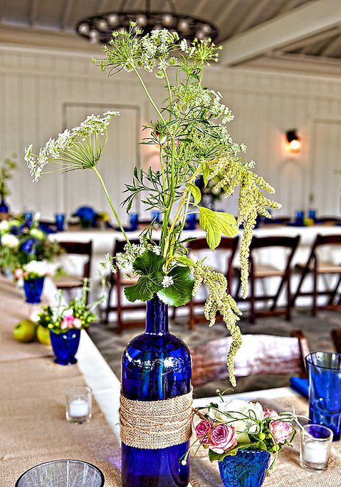 bright blue wine bottles wrapped with burlap and twine, blue vases and glasses with blooms and greenery and blue candleholders to style a rustic wedding