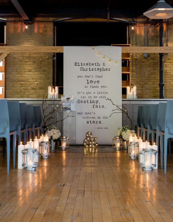 an industrial wedding ceremony space with a letter backddrop, candle lanterns, candles, branches and metal chairs for a cool look