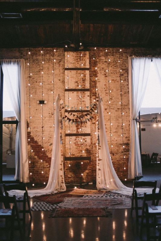 an industrial boho wedding ceremony space with shabby brick walls, lights, curtains, buntings and boho rugs for a free-spirited feel