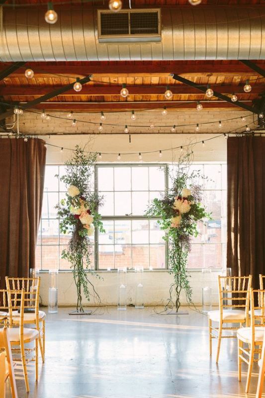 an industrial wedding ceremony space with a wooden ceiling with metal beams, lights and a lush floral altar to soften the space