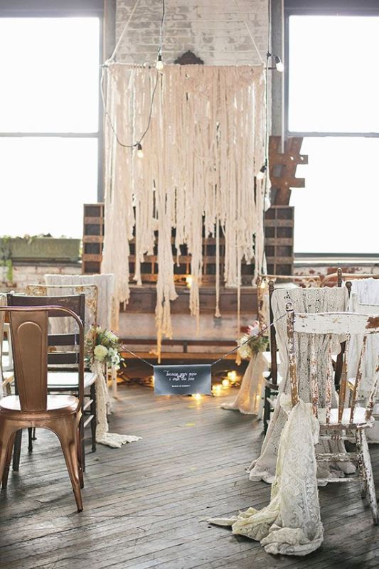 a shabby industrial wedding space with a macrame and lights backdrop, shabby chairs, blooms and a chalkboard sign