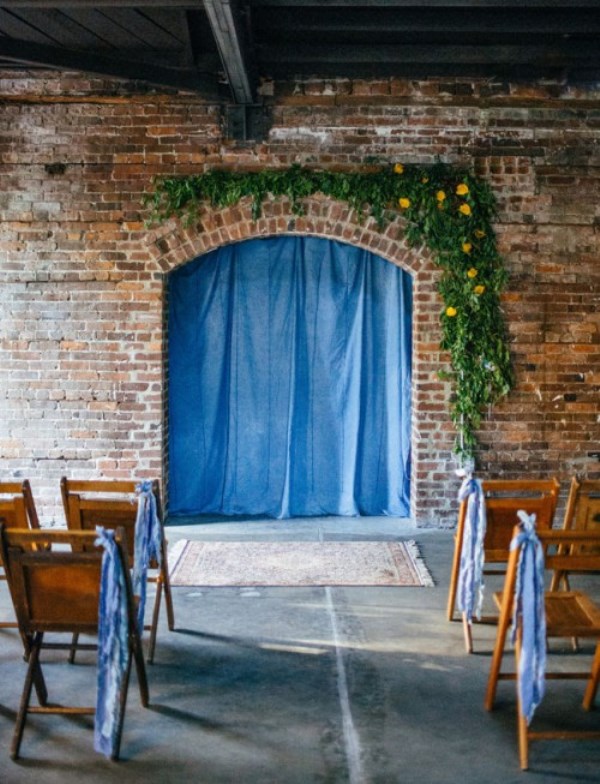 an industrial wedding space with brick walls, wooden chairs, greenery and bright bloom decor, bright blue curtains and ribbons on the chairs