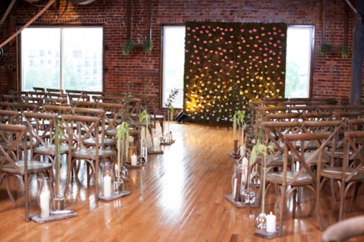 an industrial wedding ceremony space with brick walls, greenery, a greenery wall with blooms, candles and greenery attached to the chairs