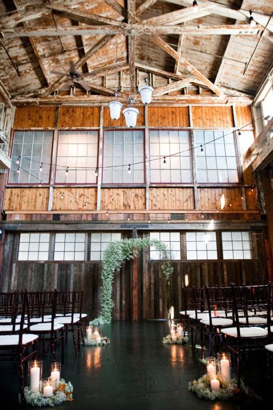 an industrial wedding ceremony space with chandeliers, lights, candles and a greeneyr wedding arch to soften the space