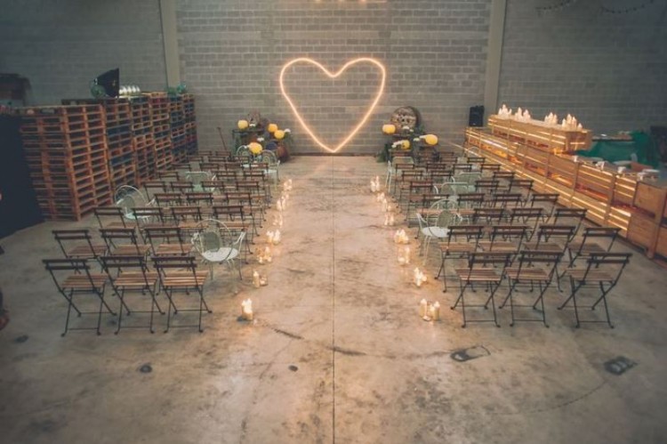 an industrial wedding ceremony space with pallets, metal and wood chairs, candles and a neon heart instead of a wedding arch