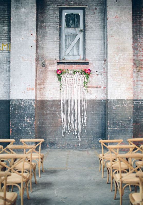 a rough industrial wedding space with a cool macrame and bright blooms and greenery backdrop plus wooden chairs