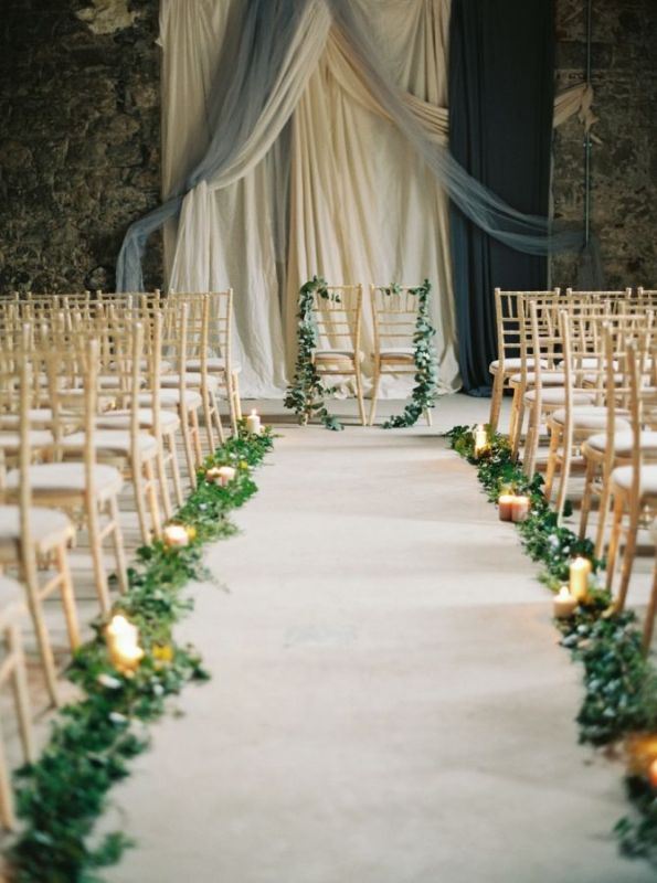 a romantic wedding backdrop of various textiles, a couple of chairs with greenery, greenery and candles lining up the aisle