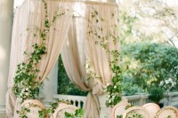 a refined and neutral wedding altar of neutral curtains, greenery and blooms decorating them and neutral chairs interwoven with greenery