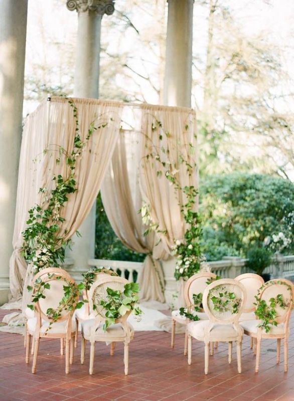 a refined and neutral wedding altar of neutral curtains, greenery and blooms decorating them and neutral chairs interwoven with greenery