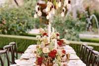 a pretty outdoor dining space with bulbs over the table and lush red and blush florals on it, red and gold glasses and chic candles