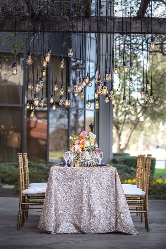 an elegant outdoor wedding reception with a silver sequin tablecloth, bright blooms and bulbs hanging over the table is amazing