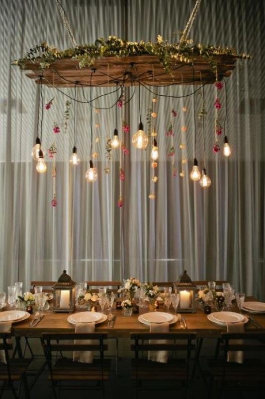 a bold wedding reception space with a wooden table, neutral blooms and an overhead installation with hanging blooms and bulbs is very elegant and rather cozy