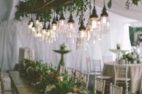 a pretty wedding reception space with lots of greenery on the table and an overhead installation with lots of greenery and bulbs as an addition to the centerpieces