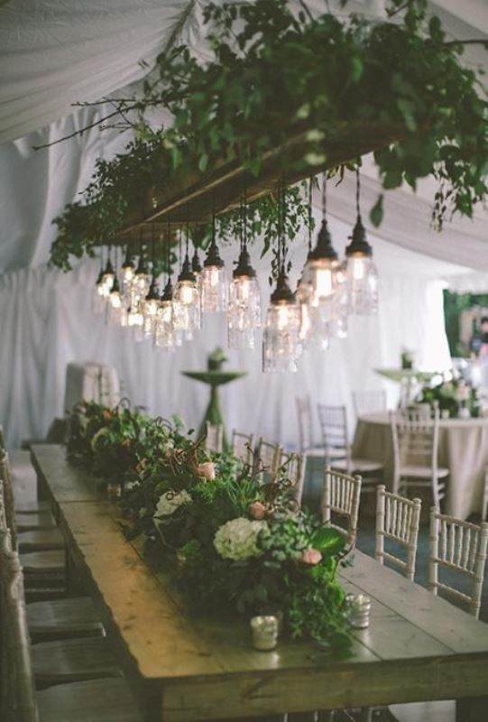 a pretty wedding reception space with lots of greenery on the table and an overhead installation with lots of greenery and bulbs as an addition to the centerpieces