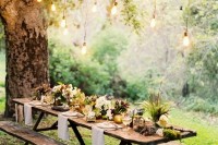 a boho outdoor wedding reception with a trestle wooden table and benches, greenery and floral arrangements and bulbs hanging down to make it more welcoming