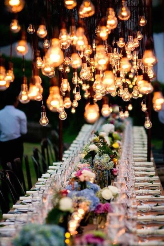 an outdoor wedding reception with lots of blooms, candles and hanging bulbs over the table to make the reception space cozier and more welcoming