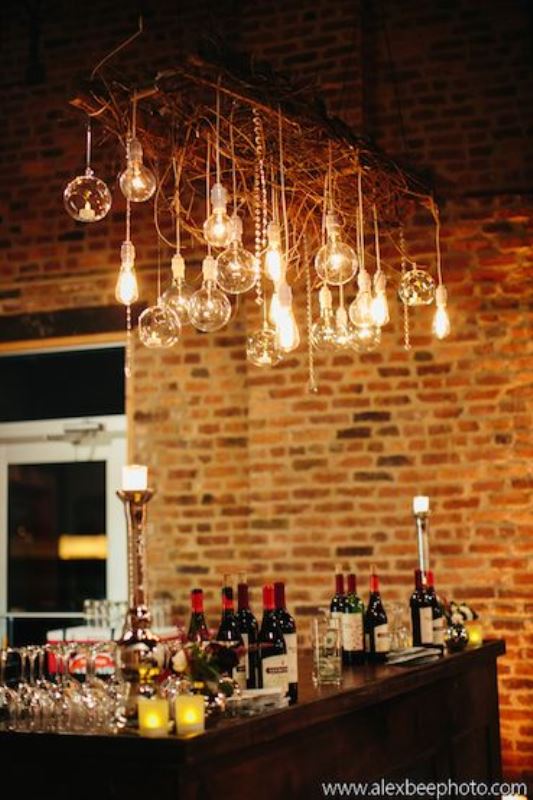 a wedding bar with candles, lots of bottles and glasses and an overhead installation of branches and lots of bulbs hanging down is all cool