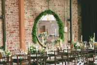 an industrial wedding venue with brick walls, a greenery garland and a mirror covered in greenery and greenery and white bloom centerpieces
