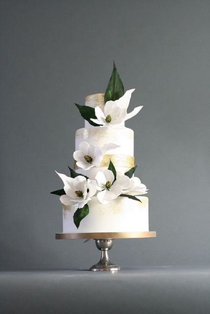 a white wedding cake with gold ribbons and white magnolias is a chic and glam idea for a modern wedding