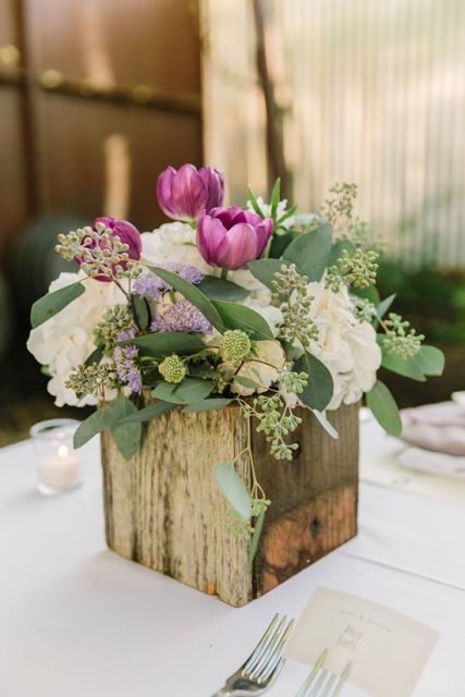 a rustic wedding centerpiece of white hydrangeas, purple tulips, greenery in a wooden cube vase