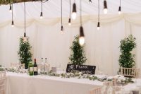a beautiful and fresh wedding reception space done in neutrals and with lots of greenery, with bulbs hanging over the tables instead of candles
