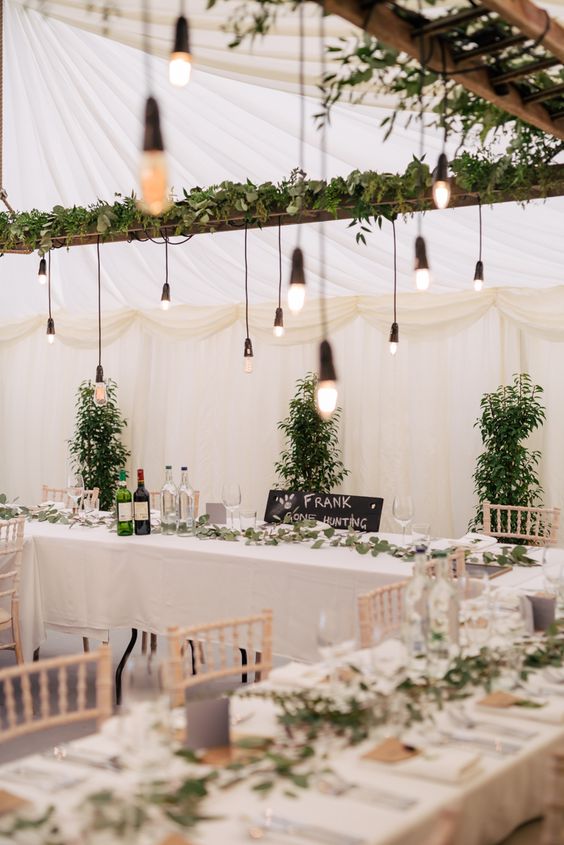 a beautiful and fresh wedding reception space done in neutrals and with lots of greenery, with bulbs hanging over the tables instead of candles