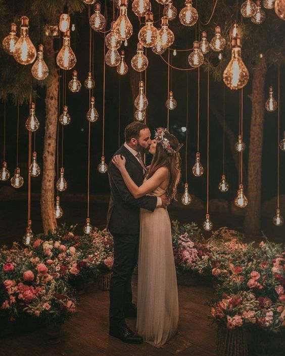 a beautiful and romantic wedding ceremony space with lots of bulbs hanging down and lush pink florals and greenery aorund