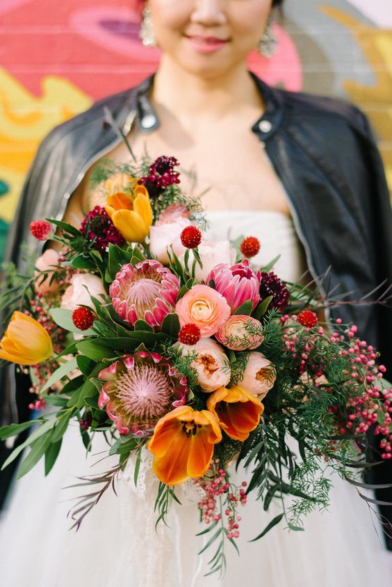 a bold and colorful wedding bouquet of orange tulips, pink king proteas and ranunculus, greenery and berries is a lovely idea for a bold wedding