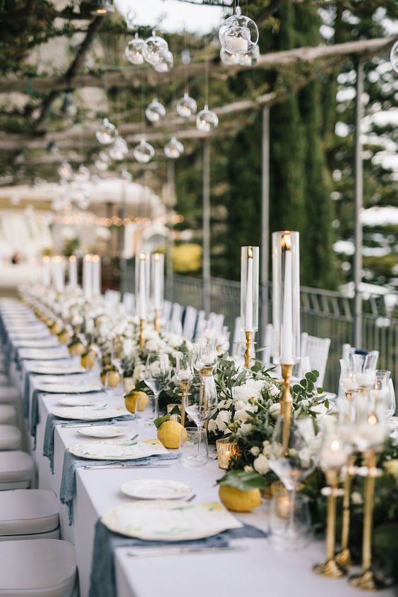 a chic wedding tablescape with a lush greenery and white bloom runner, tall and thin candles and lemons on the table